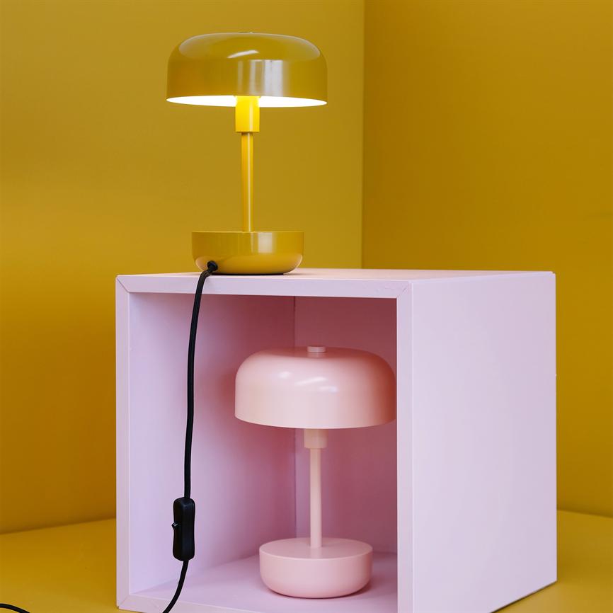 A yellow table lamp shines above a pink open box with a pink table lamp inside; black cord with switch hangs down in front of a yellow wall and table.