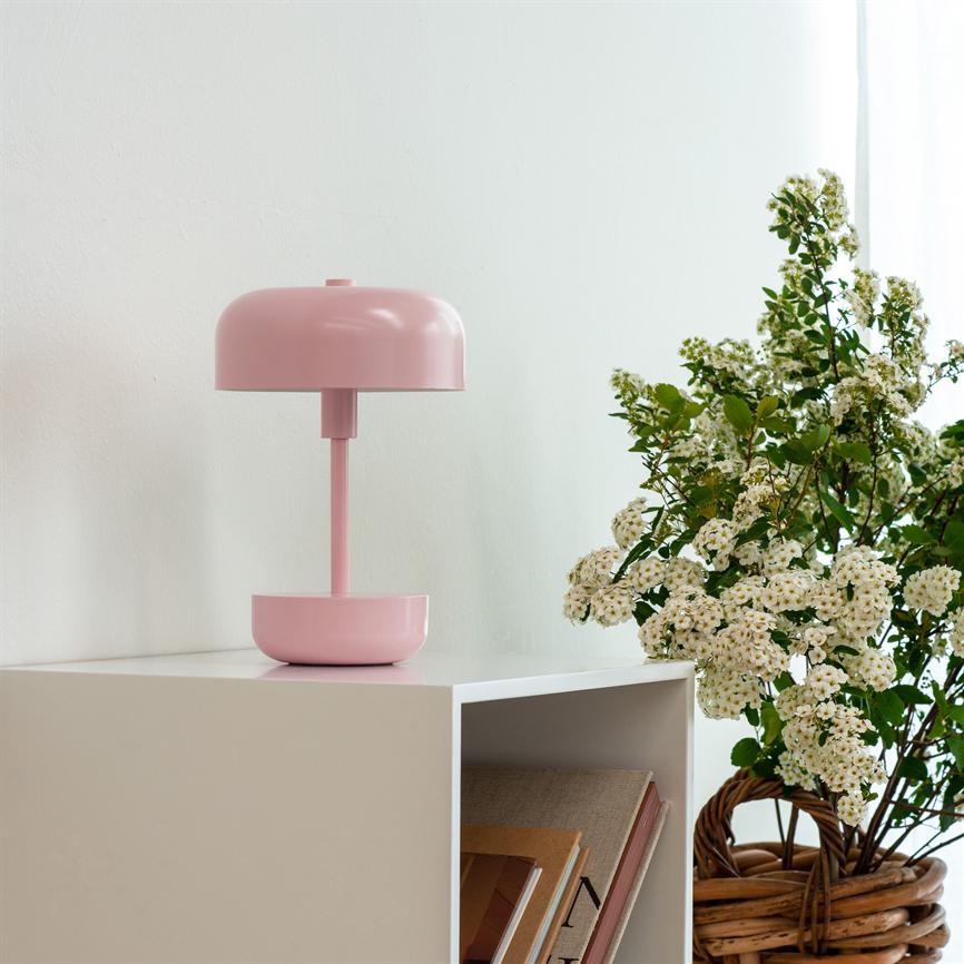 A pink table lamp stands on a white bedside table; next to it is a basket of white flowers, and an open shelving unit with stacks of books in a light, neutral room.