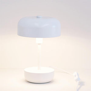 White table lamp lights dimly, with round shade and cylindrical base; it stands on a white surface in front of a neutral, light wall, with a visible power cord and switch.