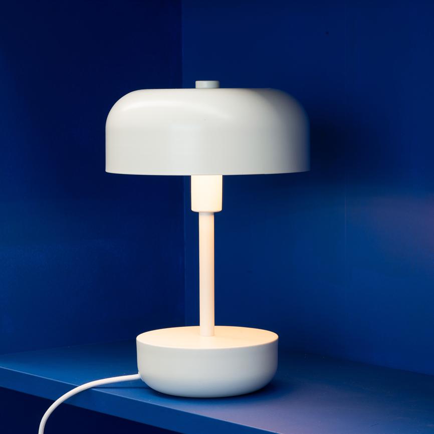 White, round table lamp emits soft, warm light; it stands on a blue shelf in front of a deep blue wall.