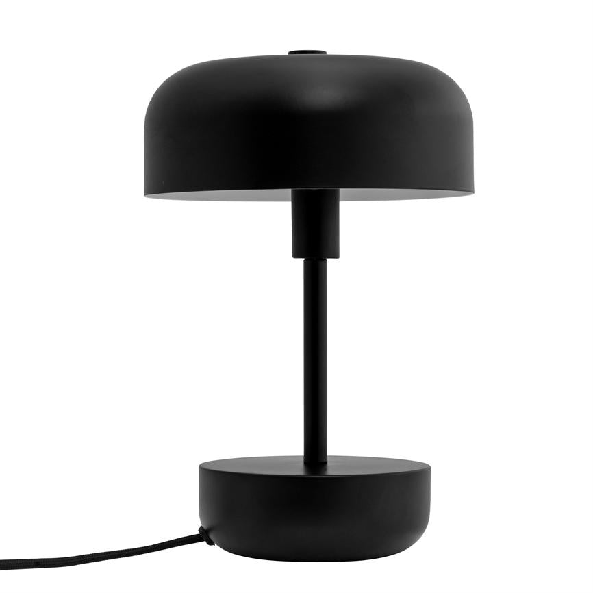 Black table lamp with round shade and cylindrical stand, switched off; round base rests on white background, with a black cloth-covered cord to the left.