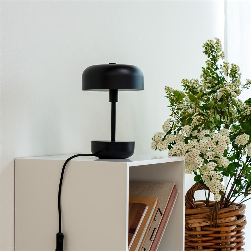 Black table lamp stands on a white open cabinet; a black cord hangs down, inside the shelf are a few books, next to it is a wicker basket with white flowers.