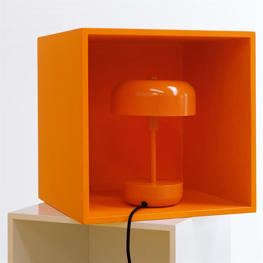 Orange table lamp stands inside an open orange cube, with black cord hanging out in front; the cube is stacked on a white cube against a white background.