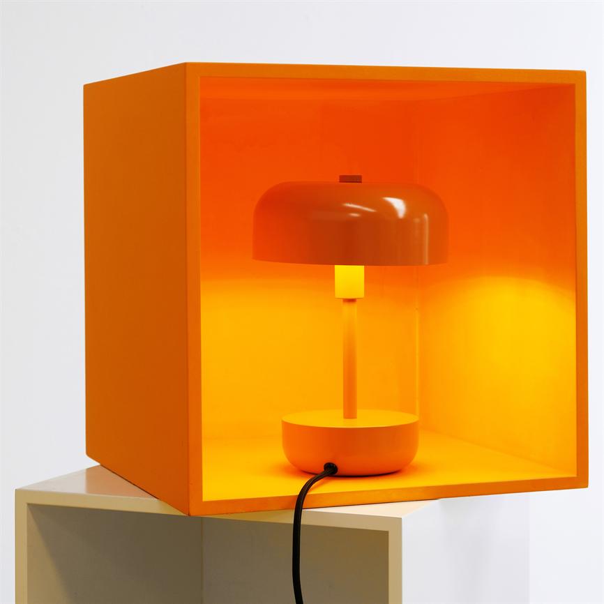 An orange table lamp shines inside an open orange box, placed on a white shelf; a black wire hangs out in front of the box.