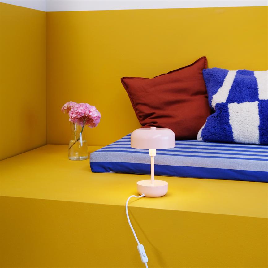 A pink table lamp is lit, casting warm light on a yellow platform; behind it is a blue-white striped mattress with a red pillowcase and a glass vase with pink flowers.
