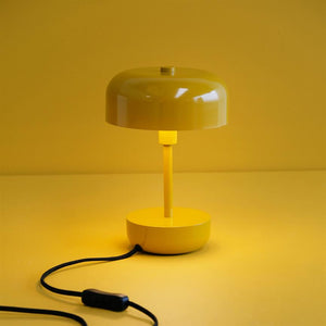 Yellow table lamp glows softly and casts warm light downwards; standing on yellow surface in front of a yellow wall, with a black cord and switch lying in front.