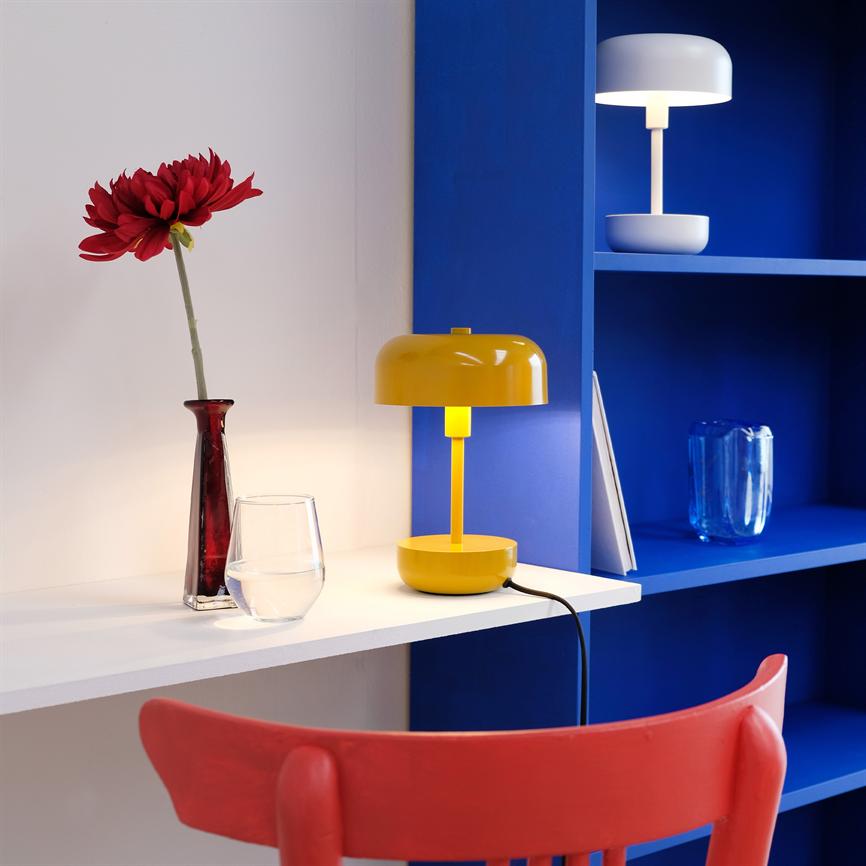 Yellow table lamp shines on a white shelf; next to a red flower in a narrow vase, in front of blue bookshelves and a red chair.