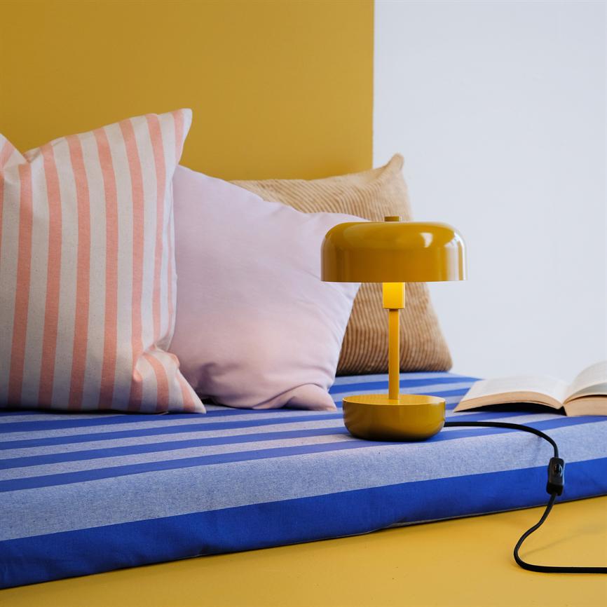 A yellow table lamp stands on a blue and light blue striped cushion next to an open book; around it are pink, striped and beige cushions in front of a yellow and white background.