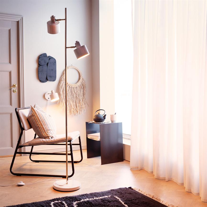 A floor lamp casts warm light over an upholstered chair with a striped cushion in a bright, cozy corner by a window with transparent curtains, a side table and a teapot.