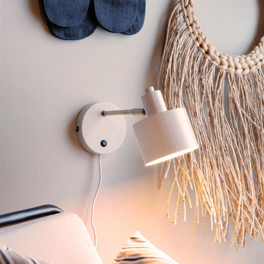A white wall lamp points downwards and emits warm, soft light; mounted above a sofa with a striped cushion, exposed wire against a light gray wall and next to a macramé wall hanging.
