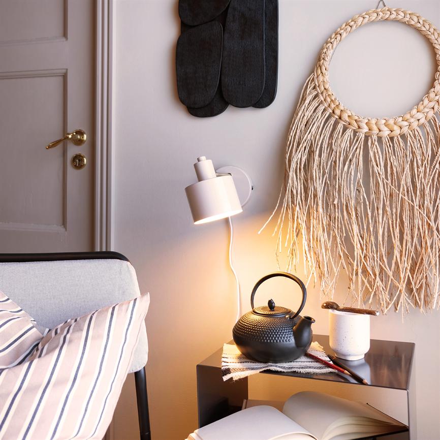 A black cast-iron teapot stands on a small table, lit by a wall lamp; next to a cup, open book and wicker wall decoration in a cozy corner of the living room.