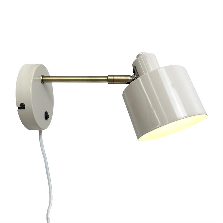 Wall lamp with white metal shade and round white wall base, mounted on a short brass arm; the screen is slightly downwards and lit, with a white cloth-covered cord hanging.