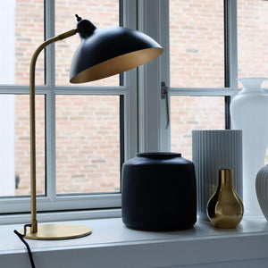 Black and brass desk lamp bends over vases on a white windowsill; arrangement includes a large matte black vase, ribbed white vase and small gold colored vase; brick facade seen outside.