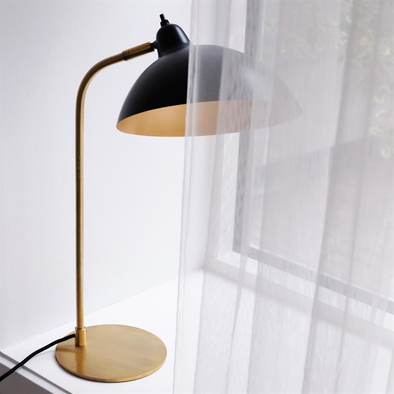 A black and gold table lamp stands on a white windowsill, partially covered by a light white curtain, with curved brass stand and black shade with golden interior.