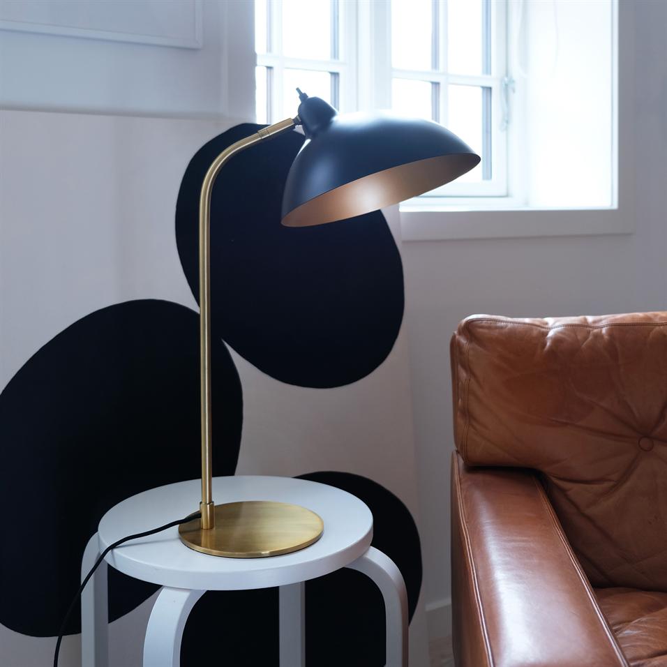 A black table lamp with a golden base and angled shade stands on a round white side table next to a light brown leather sofa in front of a window and a wall with large black circles.