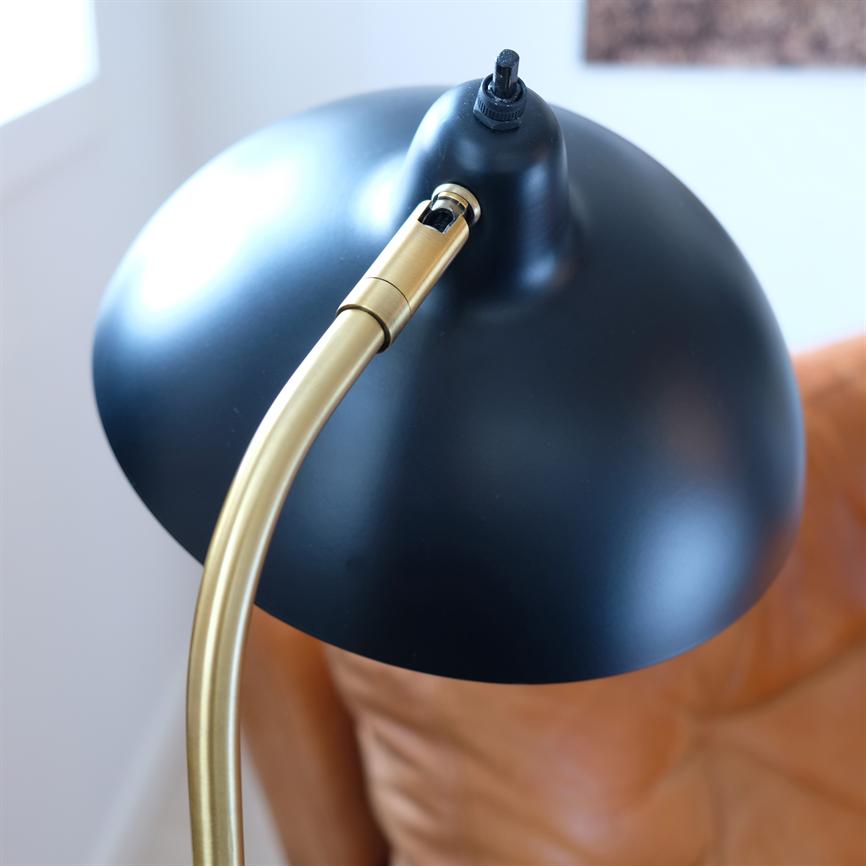 Black round lampshade on a bendable brass arm, angled downward over a brown leather upholstered seat in a bright room by a window.