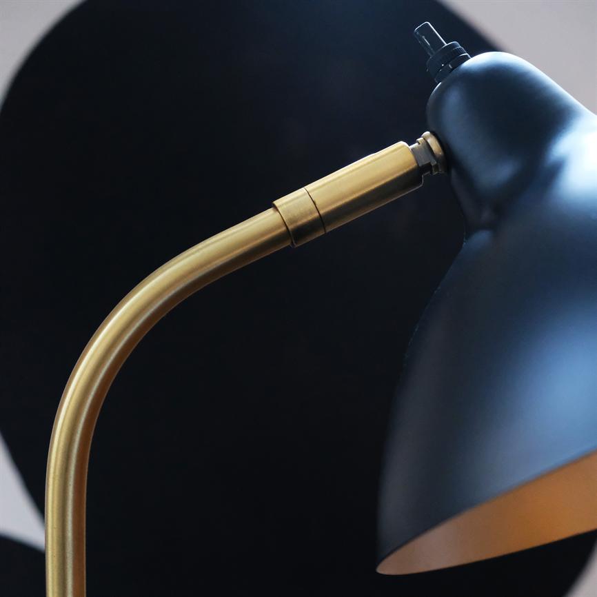 Desk lamp with black shade and curved, brass-colored arm points downwards in front of a dark, circular background in a minimalist interior.