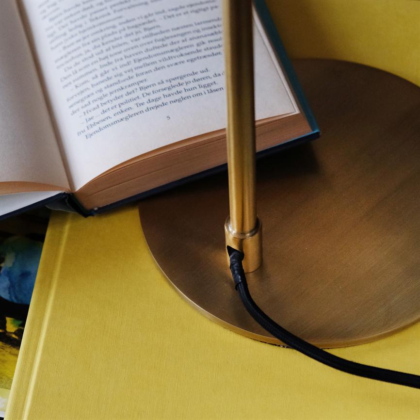 A table lamp stands on a round brass base; its black cord runs over a yellow table next to an open book with printed Danish text.
 
 Transcription of visible text (partially illegible):
 "He had been in bed for three days
 The estate agent turned the key in the lock
 5"
 
 Other text on the page is not fully legible in the image.