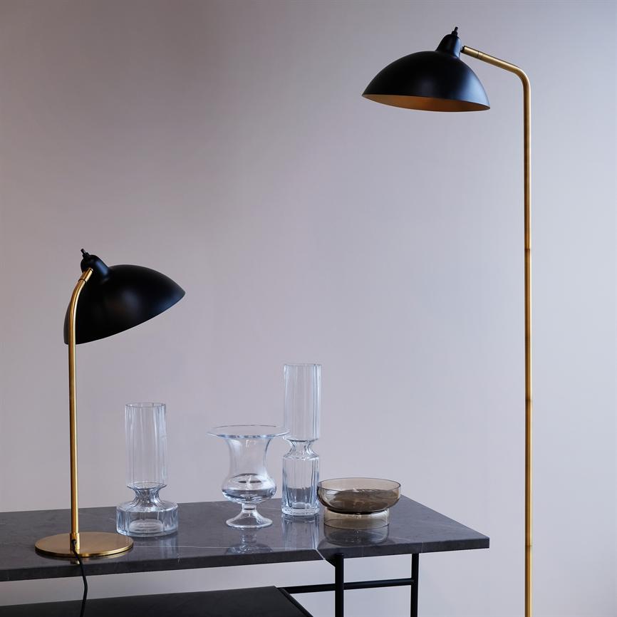 A table lamp and a floor lamp in black and brass bend their shades down over a dark marble table, where several clear glass vases and a smoked glass bowl stand in front of a light, empty wall.