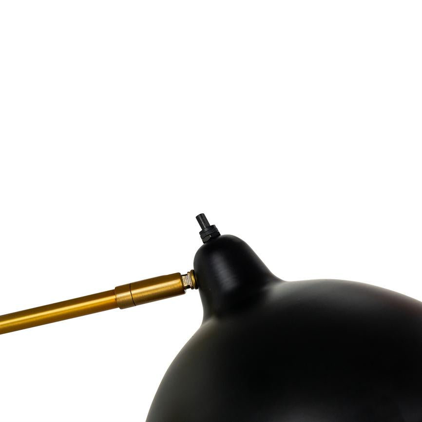 Black metal shade of a desk lamp, connected to a gold colored arm with visible hinge and adjustable connection, leans in the foreground against a uniform white background.