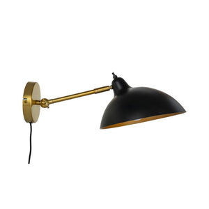 Wall lamp with black dome shade and gold-coloured metal arm, mounted on a round wall bracket; the arm is extended and the screen tilted downwards against a white, neutral background.