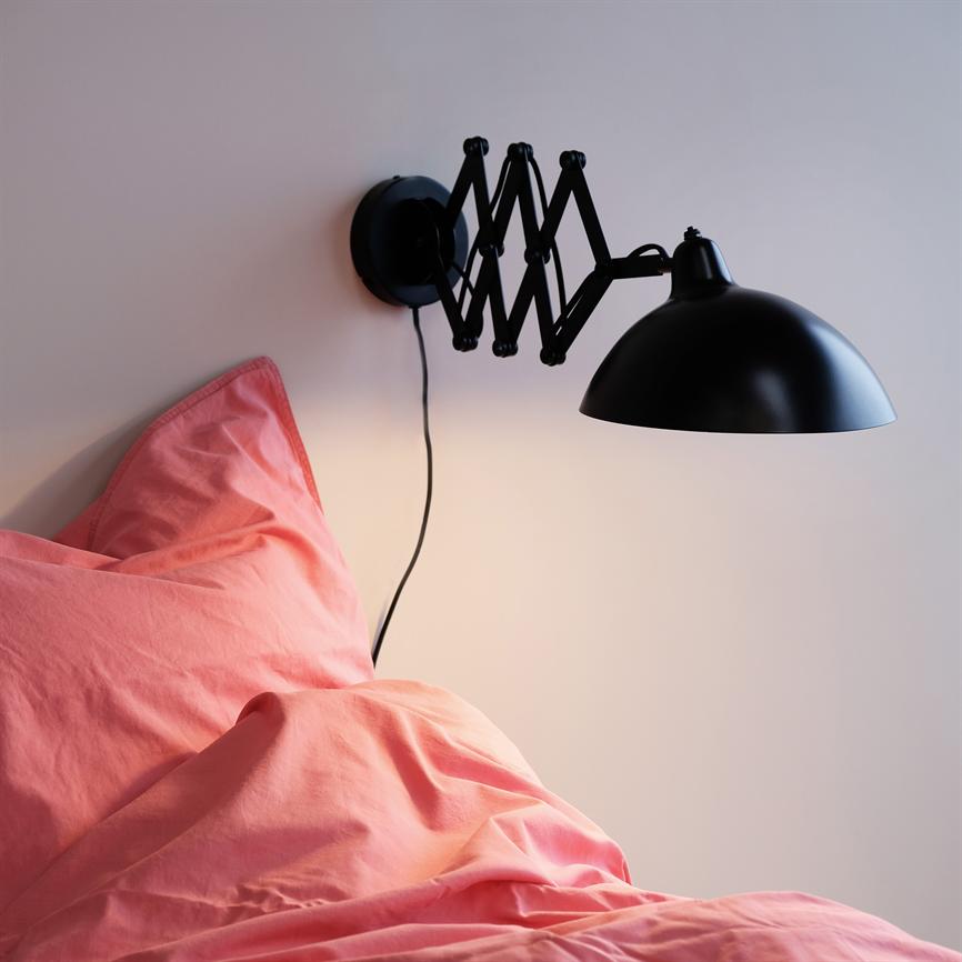 Black wall lamp with scissor-jointed arm, extended and shining downwards; above a bed with coral/pink duvet cover and pillow, mounted on a light wall.