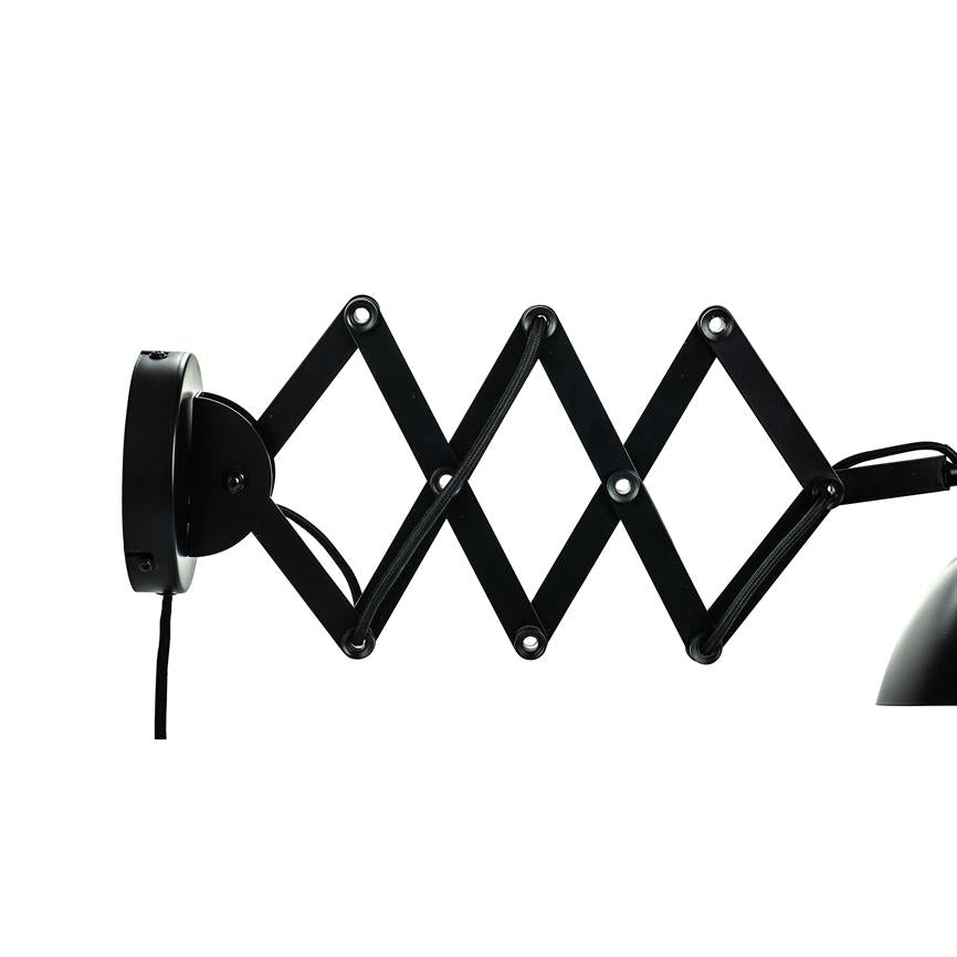 Black wall lamp with scissor arm extended in zigzag; the arms unfold from a round wall bracket, cable hangs down, and a partial lampshade is seen to the right against a white background.