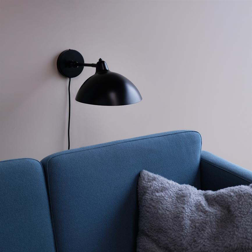 A black wall lamp hangs and points down above a blue sofa; the sofa's backrest is flanked by a soft light gray plush cushion against a light beige wall.
