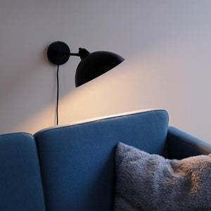 A black wall lamp casts warm light over the back of a blue sofa, with a gray soft cushion on the seat in a calm, neutral living room environment.