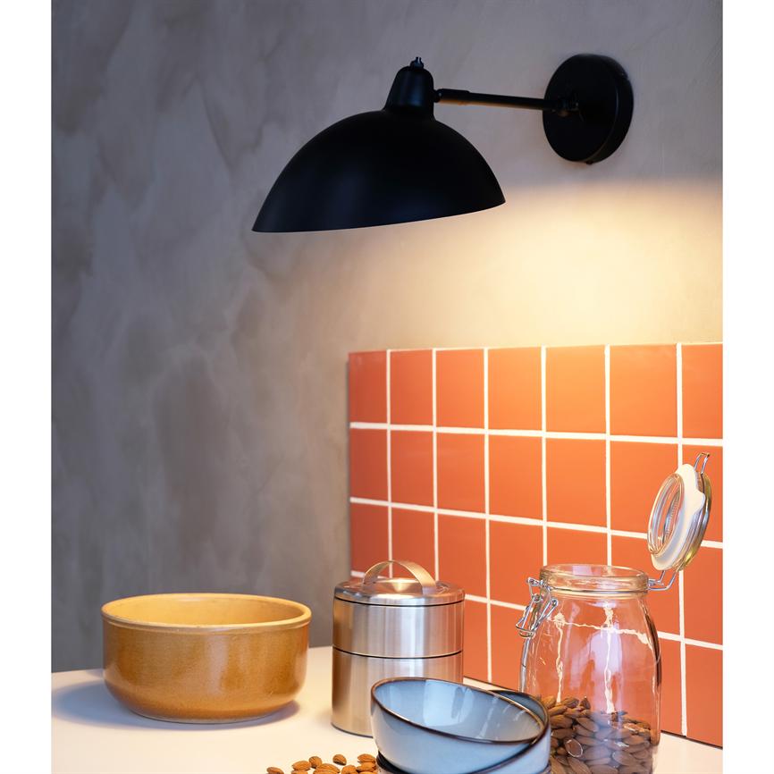 A black wall lamp shines down on a red tile back panel and a kitchen table with a ceramic bowl, metal container, open glass jar and scattered almonds.