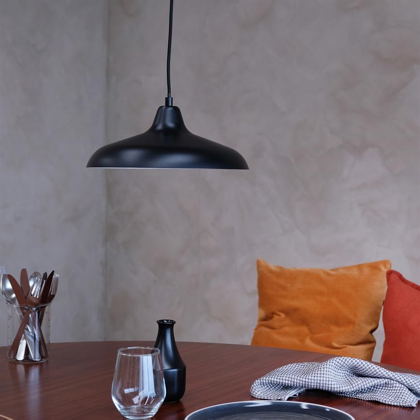 Black hanging lamp hangs above a round wooden table with a glass, a small black vase, cutlery in a glass, a checkered napkin and orange cushions in front of a light textured wall.