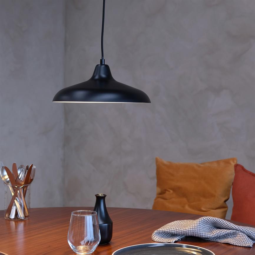 A black ceiling lamp hangs above a wooden table and casts soft light on drinking glasses, a small black vase and glass cutlery in a simple dining area with cushions against a raw wall.