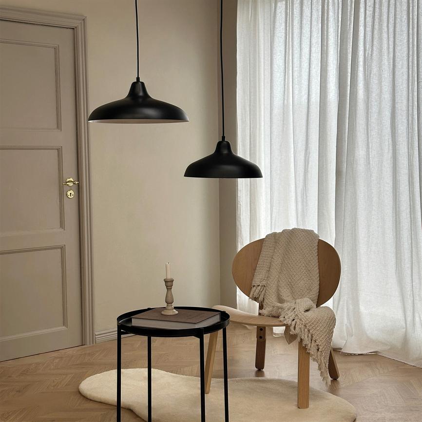 A wooden chair carries a beige woven rug, placed by a round black side table with a single candle holder; two black pendant lamps hang above the chair, in front of light curtains and a wooden floor.