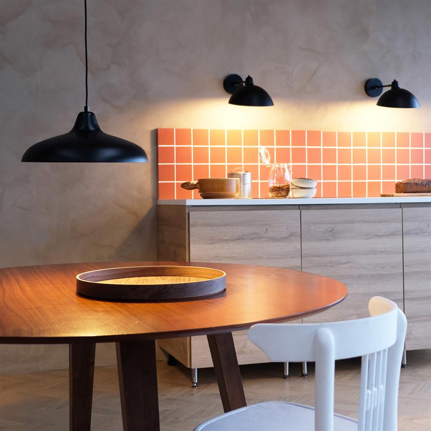 Round wooden table stands in the foreground with a round tray; a white chair next to it; behind a kitchen bench with light wooden cabinets, orange tiles and three black lamps.