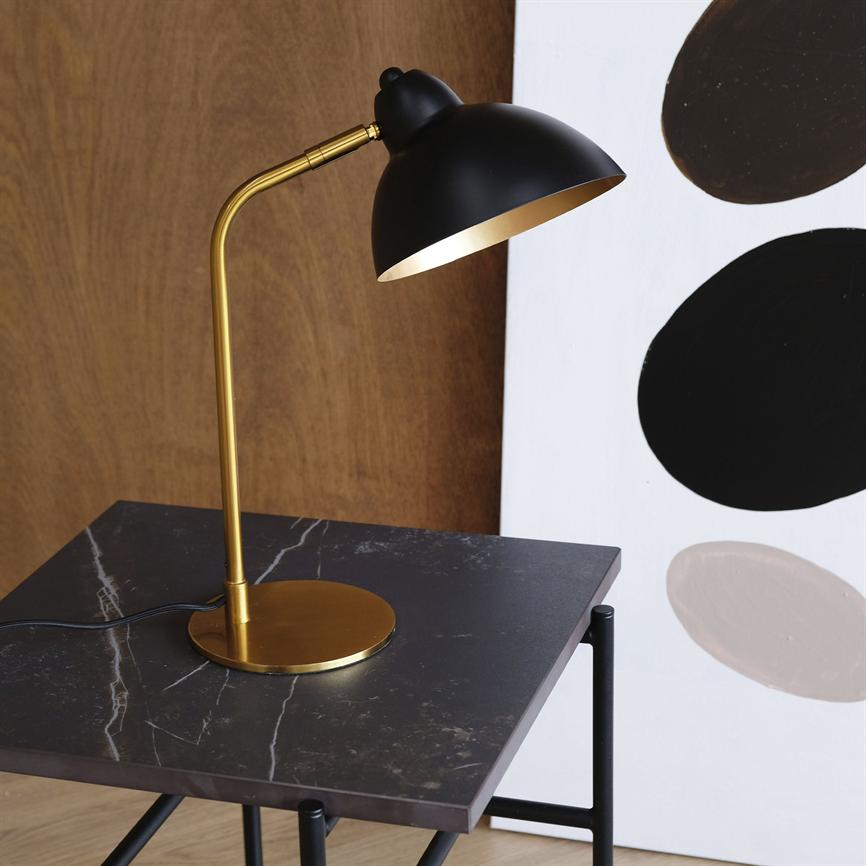 Table lamp with black shade and golden rod shines downwards, stands on a dark marble table in front of a wood-clad wall and a white painting with three large black/beige ovals.
