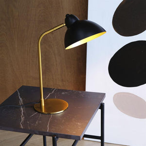 Object: Black table lamp with golden rod and foot; Action: illuminates and casts warm light downwards; Context: on dark marble table in front of wooden wall and a canvas with black and beige circles.