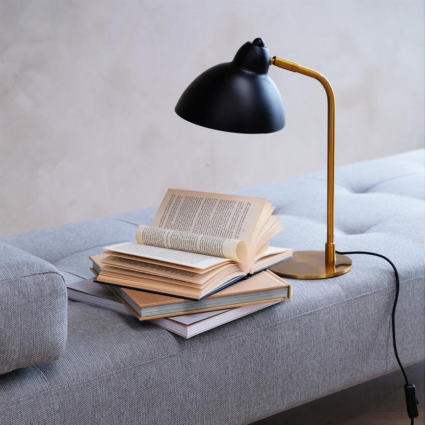 A black and gold reading lamp illuminates an open book lying on a stack of books on a gray sofa in a bright, minimalist room; no readable text to transcribe.