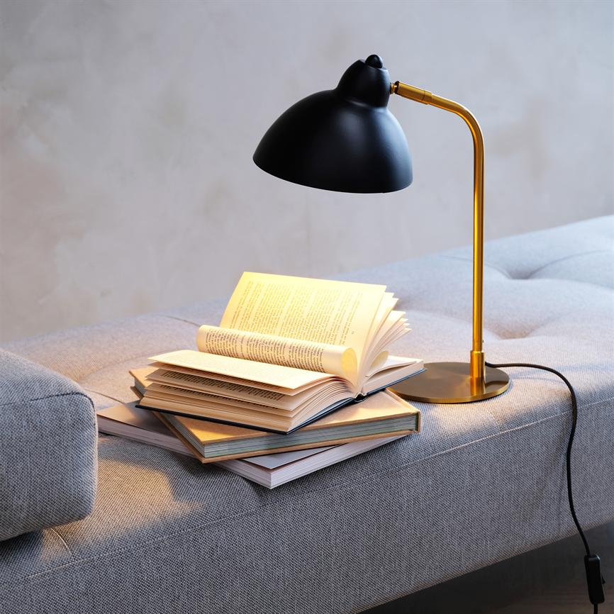 A black and gold table lamp shines over a stack of open books lying on a light gray sofa in a calm, neutral room.
