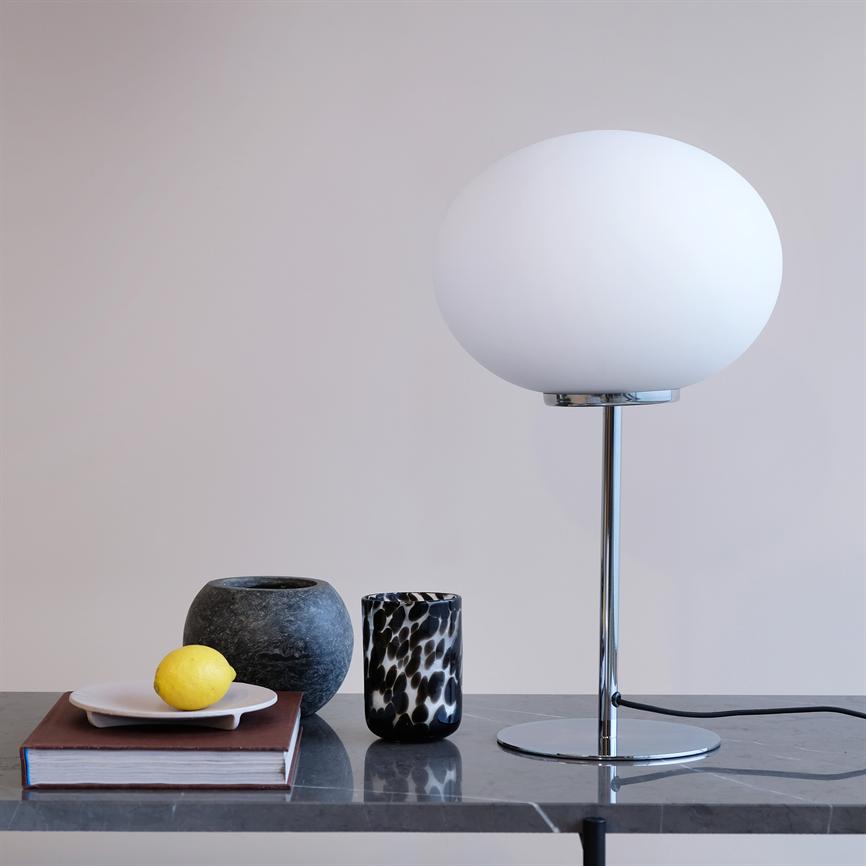 A white round table lamp stands on a chrome stand on a dark marble tabletop; next to a book with a plate and lemon and two vases in front of a beige wall.