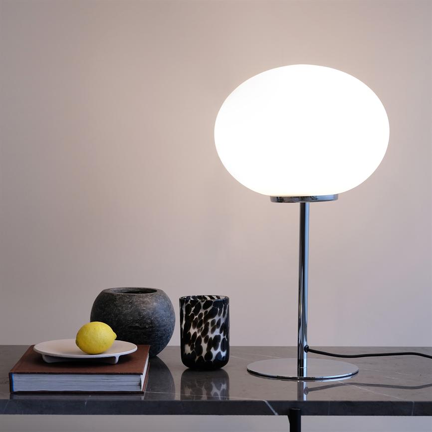 Table lamp with round, white glass shade shines softly on a marble table, flanked by a book with a plate and lemon, a dark stone urn and a black and white patterned glass.