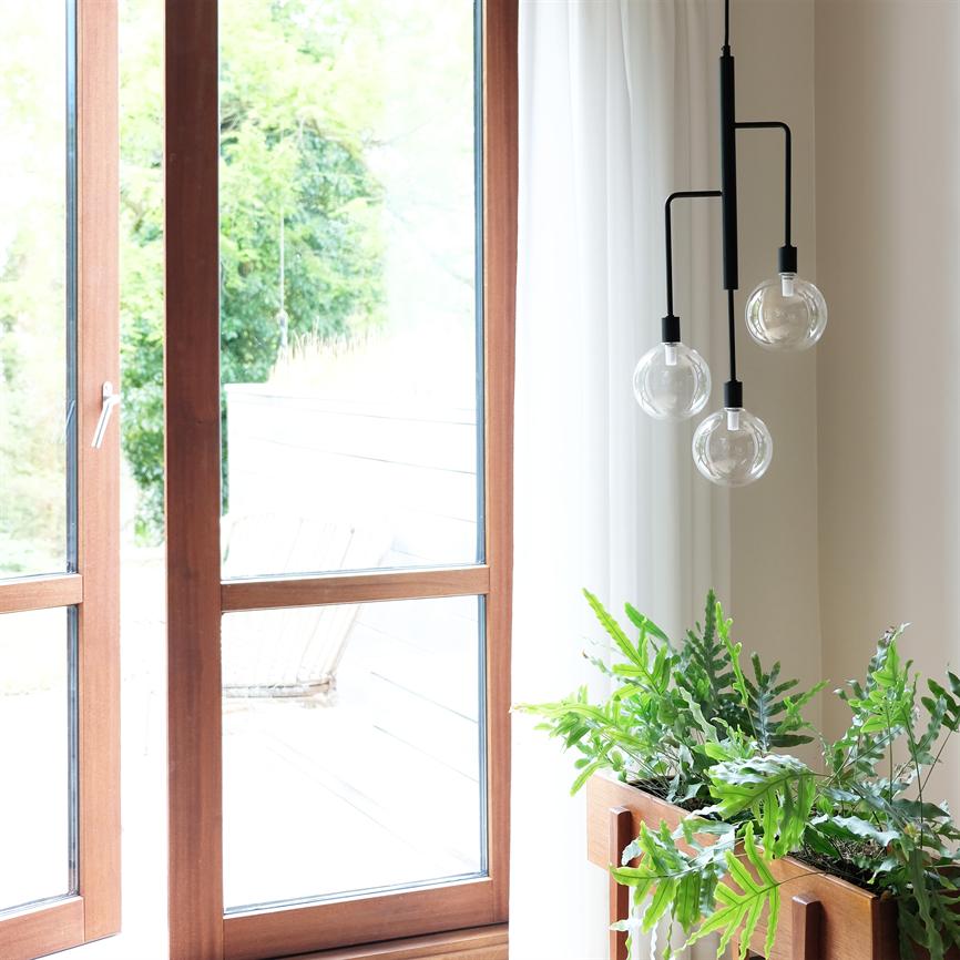 A black three-armed pendant hangs with three bright light bulbs by a large wooden window; sunlight falls on a wooden pot with lush green houseplants on the windowsill.