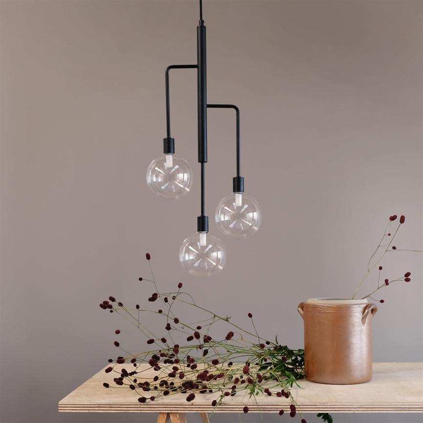 A modern one pendant with three clear glass domes hanging over a table where wild maroon flowers are scattered, and a brown ceramic jar stands against a dusty pink wall.