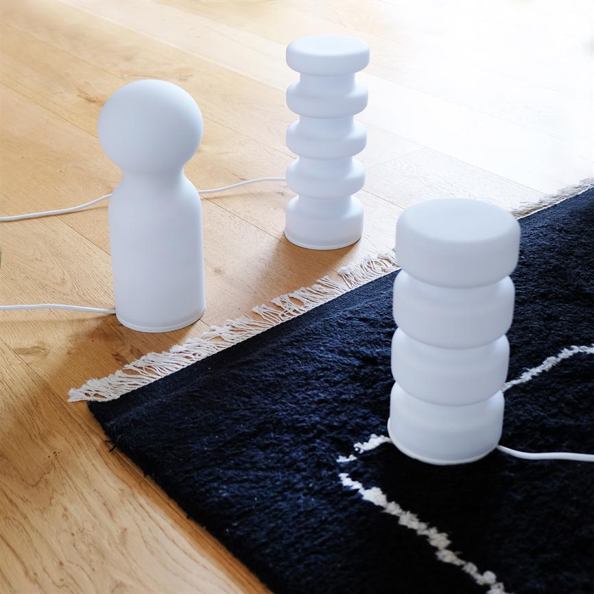 Three white table lamps are placed on a light wooden floor at the edge of a dark carpet with white fringes; the lamps' rounded shapes and visible electric cables are clearly visible.