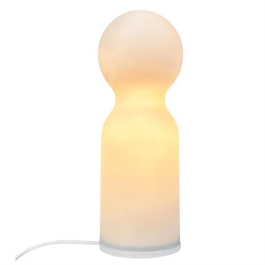Lamp emits a warm golden glow from a round top and cylindrical body, stands on white background with white cord visible at base.