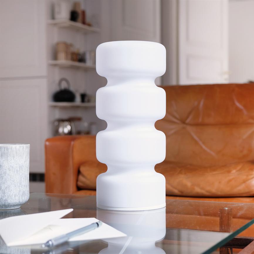 White, wavy table lamp stands on a glass table; surrounded by a notepad and pen in the foreground, in front of a brown leather sofa and shelves in a cozy living room.