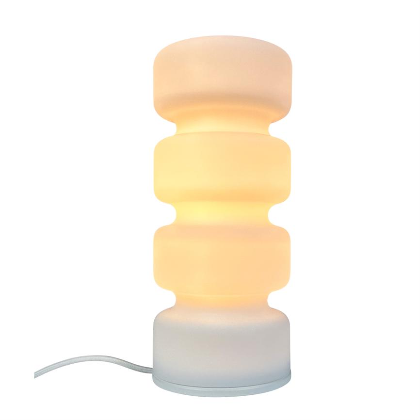 A white, curved table lamp emits warm, diffused yellow light; four close-fitting round segments stand vertically on a round foot with a white fabric cord to left on white background.