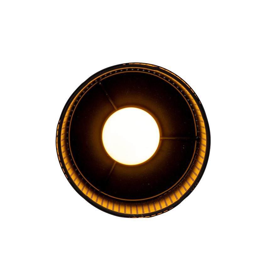 A round ceiling lamp emits intense warm light from the centre, surrounded by a dark, perforated inner shade, which casts a golden glow against a white background.