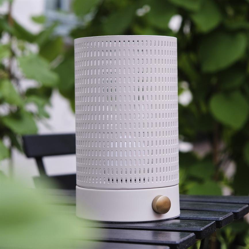 White cylindrical electronic device with perforated surface and bronze colored dial stands on a black wooden table on an outdoor terrace surrounded by green leaves.