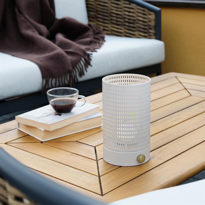 A perforated cylindrical table lamp casts warm, dim light on a wooden table; next to it is a cup of coffee on two books in front of an outdoor sofa with white cushions and a brown rug.
