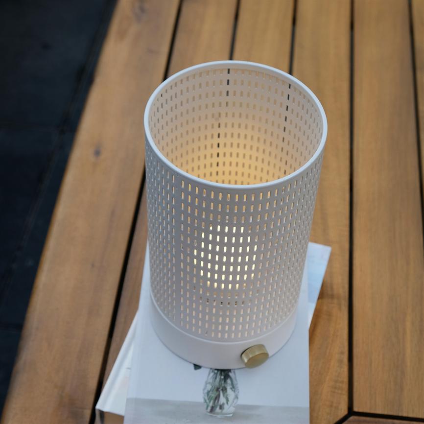 Cylindrical, white perforated table lamp emits soft, warm light; it is on a book on a wooden table in an outdoor, subdued environment.